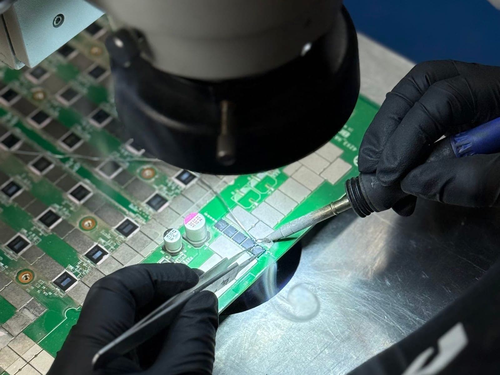 Gloved hands reworking a hashboard under a stereo microscope in the ACS repair lab